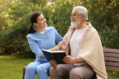 Photo of Smiling nurse with elderly man in glasses with book on bench outdoors