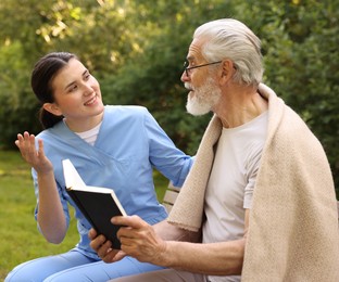 Photo of Smiling nurse with elderly man in glasses with book on bench outdoors