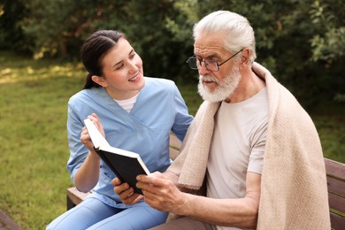 Photo of Smiling nurse with elderly man in glasses with book on bench outdoors