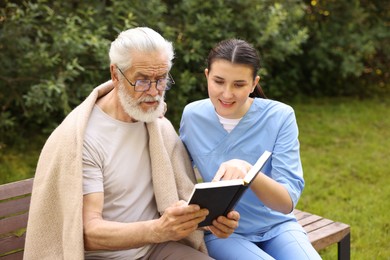 Photo of Smiling nurse with elderly man in glasses with book on bench outdoors