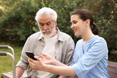 Photo of Smiling nurse and elderly man using smartphone on bench outdoors