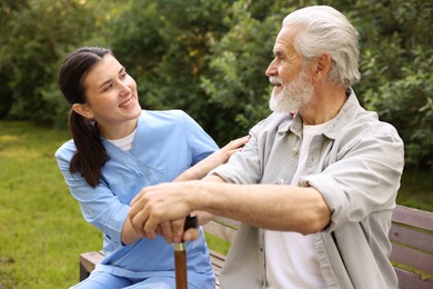 Photo of Smiling nurse supporting elderly man with walking cane on bench outdoors