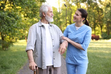 Photo of Smiling nurse supporting elderly man with walking cane outdoors