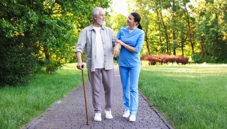 Photo of Smiling nurse supporting elderly man with walking cane outdoors