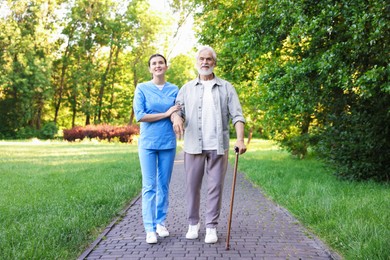 Photo of Smiling nurse supporting elderly man with walking cane outdoors