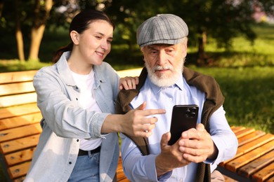 Photo of Smiling caregiver and elderly man using smartphone on bench outdoors