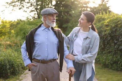 Photo of Smiling caregiver supporting elderly man with walking cane outdoors