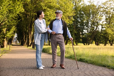 Photo of Smiling caregiver supporting elderly man with walking cane outdoors