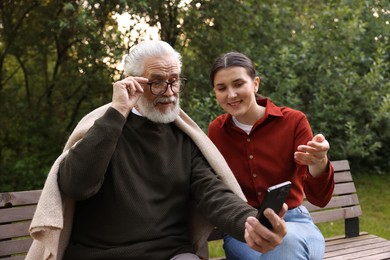 Photo of Smiling caregiver and elderly man in glasses using smartphone on bench outdoors