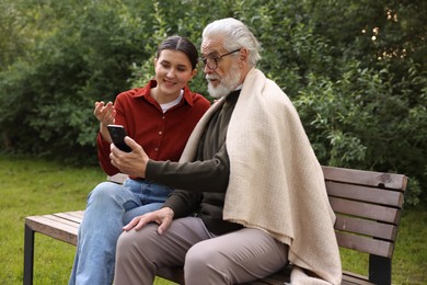 Photo of Smiling caregiver and elderly man in glasses using smartphone on bench outdoors