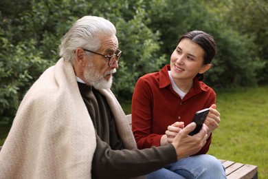 Photo of Smiling caregiver and elderly man in glasses using smartphone on bench outdoors