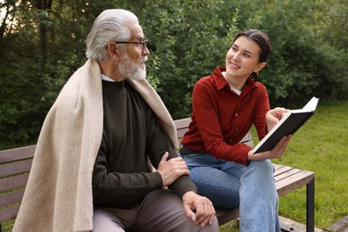 Photo of Smiling caregiver and elderly man in glasses reading book on bench outdoors