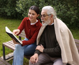 Photo of Smiling caregiver and elderly man in glasses reading book on bench outdoors