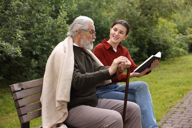 Photo of Smiling caregiver and elderly man in glasses with walking cane reading book on bench outdoors