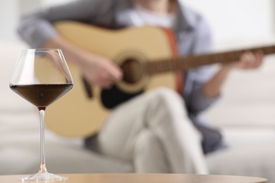 Photo of Glass with red wine on table and man playing guitar indoors, selective focus
