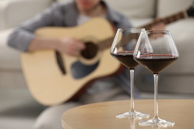 Photo of Glasses with red wine on table and man playing guitar indoors, selective focus