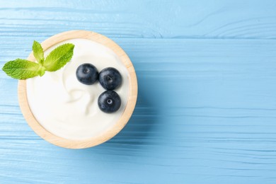 Photo of Tasty yoghurt with blueberries and mint in bowl on light blue wooden table, top view. Space for text