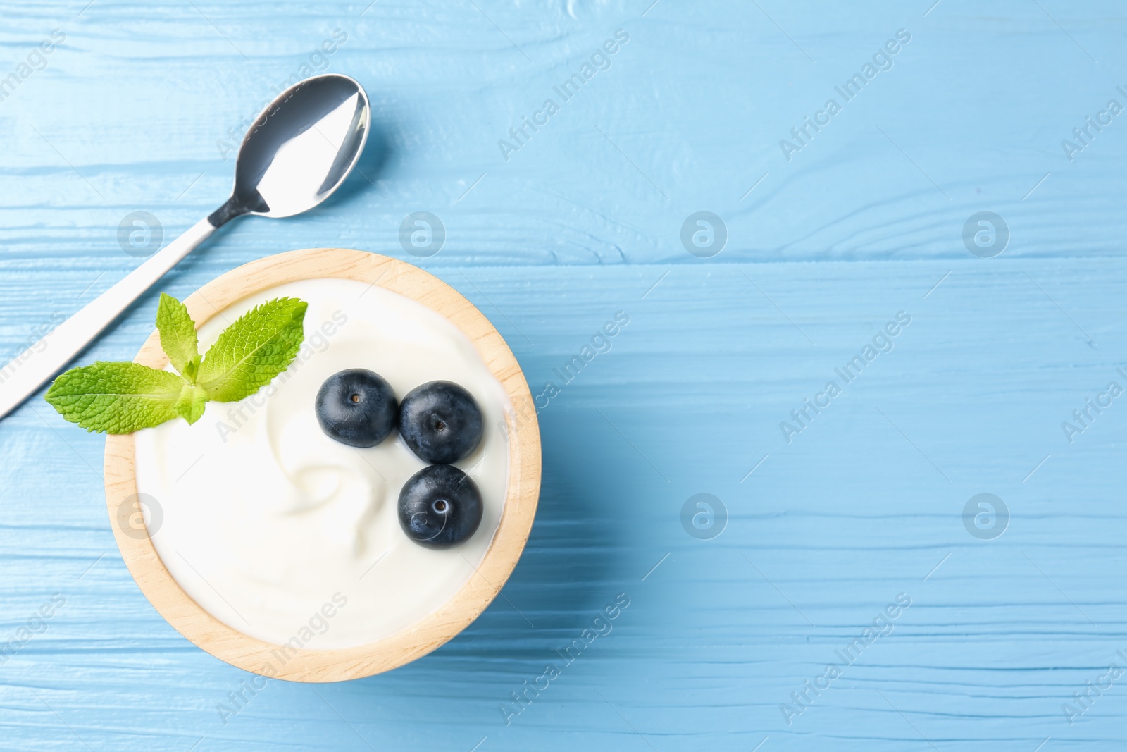 Photo of Tasty yoghurt with blueberries and mint in bowl on light blue wooden table, top view. Space for text