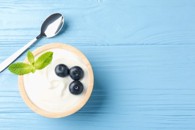 Photo of Tasty yoghurt with blueberries and mint in bowl on light blue wooden table, top view. Space for text