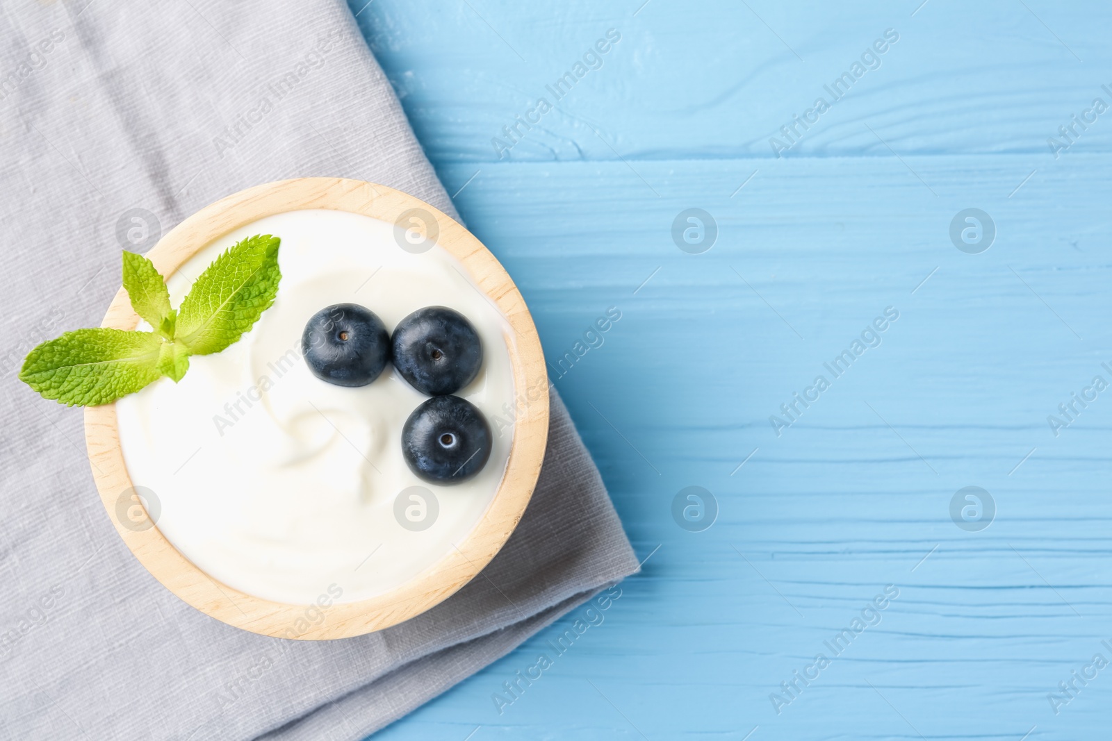 Photo of Tasty yoghurt with blueberries and mint in bowl on light blue wooden table, top view. Space for text