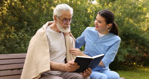 Photo of Nurse with elderly man in glasses with book on bench outdoors