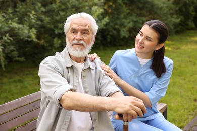Photo of Smiling nurse supporting elderly man with walking cane on bench outdoors