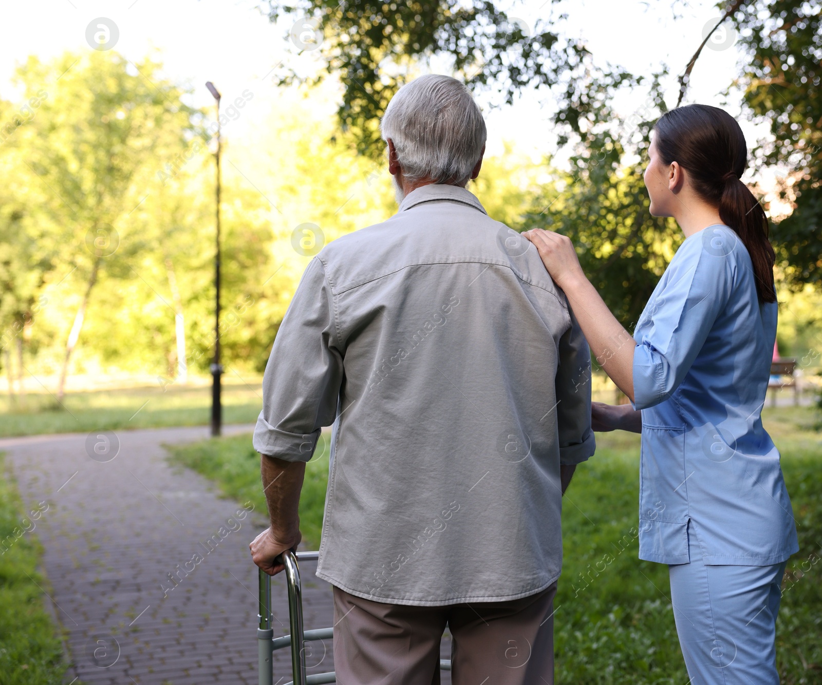 Nurse supporting elderly man who using walking frame outdoors, back view Photo of Nurse supporting elderly man who using walking frame outdoors, back view
