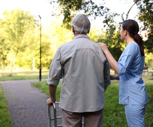 Photo of Nurse supporting elderly man who using walking frame outdoors, back view