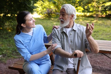 Photo of Smiling nurse supporting elderly man with walking cane on bench outdoors