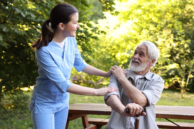 Photo of Smiling nurse supporting elderly man with walking cane outdoors
