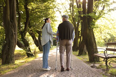 Photo of Smiling caregiver supporting elderly man who using walking frame outdoors, back view
