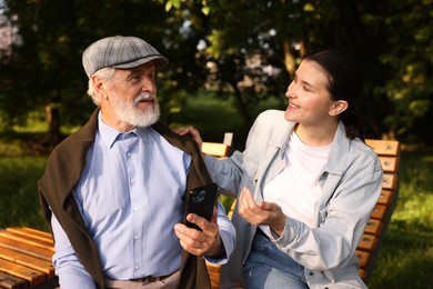Photo of Smiling caregiver and elderly man using smartphone on bench outdoors