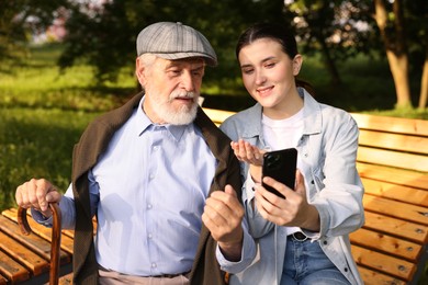 Photo of Smiling caregiver and elderly man using smartphone on bench outdoors