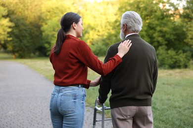 Photo of Caregiver supporting elderly man who using walking frame outdoors, back view