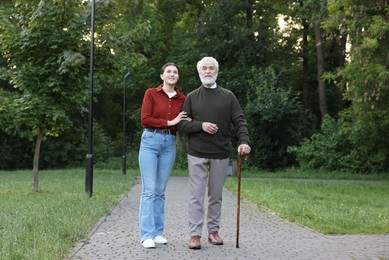 Photo of Smiling caregiver supporting elderly man who using walking cane outdoors