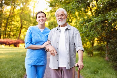 Photo of Smiling nurse supporting elderly man with walking cane outdoors