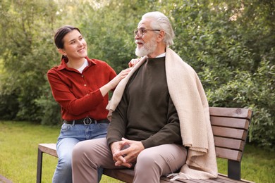 Photo of Smiling caregiver supporting elderly man in glasses on bench outdoors