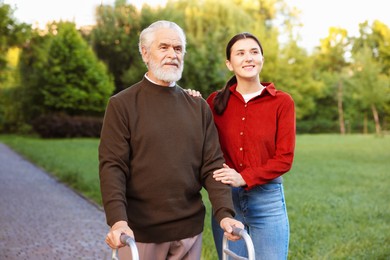 Photo of Smiling caregiver supporting elderly man who using walking frame outdoors