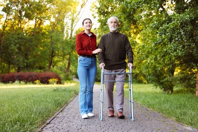 Photo of Smiling caregiver supporting elderly man who using walking frame outdoors