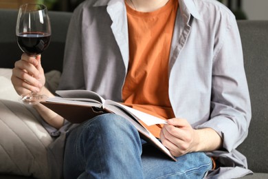 Photo of Woman with glass of wine reading book on sofa at home, closeup