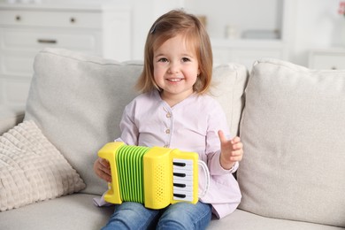 Photo of Cute little girl playing toy accordion on sofa at home