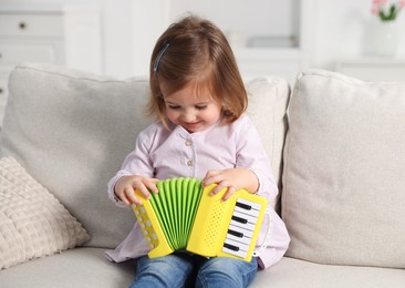 Photo of Cute little girl playing toy accordion on sofa at home