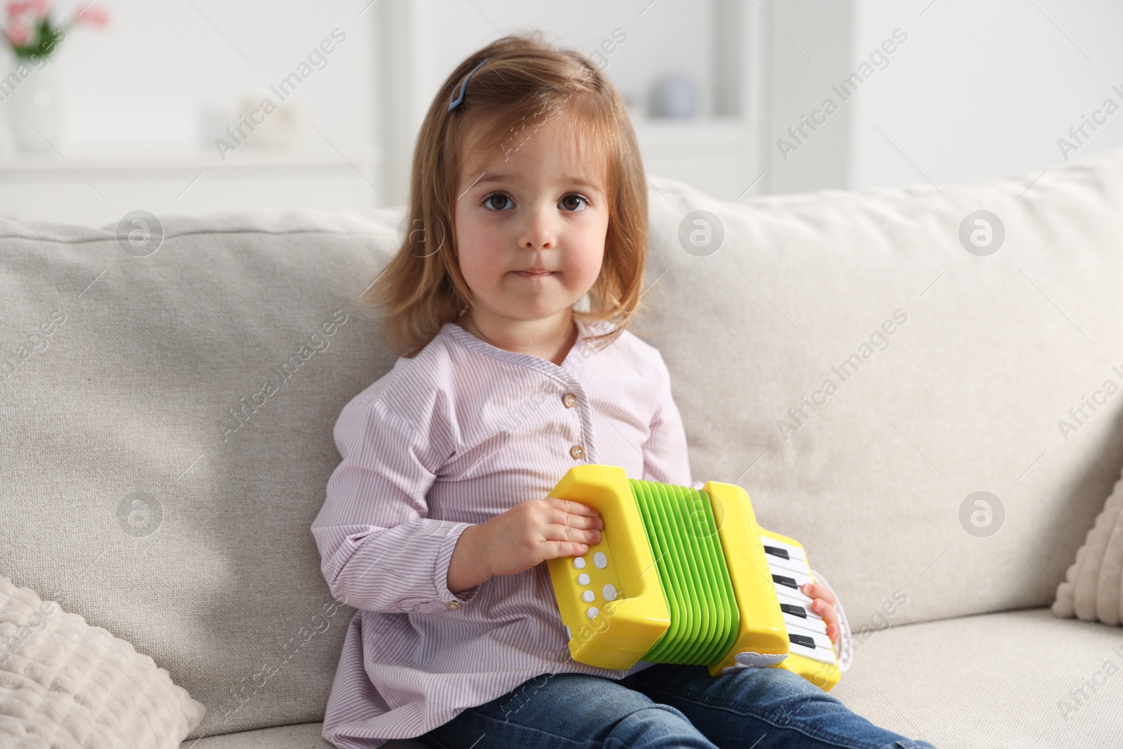 Photo of Cute little girl playing toy accordion on sofa at home