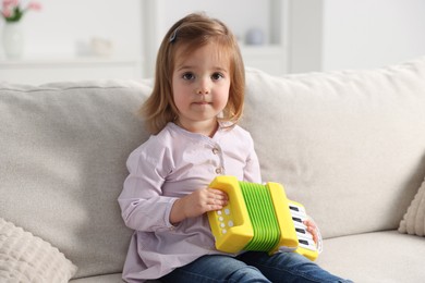 Photo of Cute little girl playing toy accordion on sofa at home
