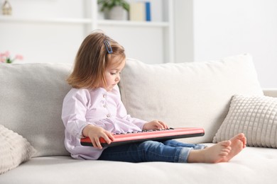 Photo of Cute little girl playing toy electric piano on sofa at home
