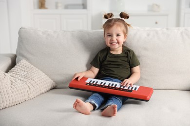 Photo of Cute little girl playing toy electric piano on sofa at home