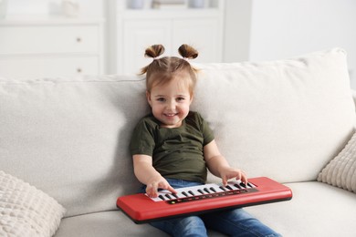 Photo of Cute little girl playing toy electric piano on sofa at home