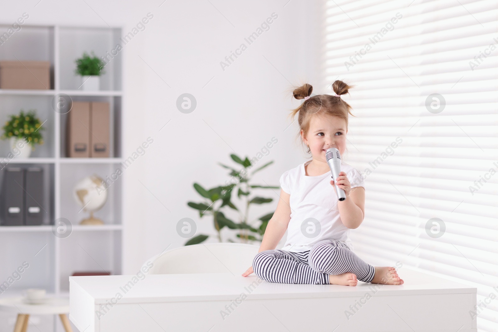 Cute little girl with microphone on white table at home, space for text Photo of Cute little girl with microphone on white table at home, space for text