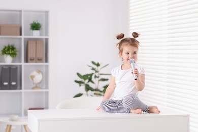 Photo of Cute little girl with microphone on white table at home, space for text
