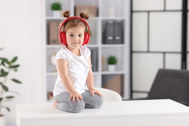 Photo of Cute little girl in headphones on white table at home, space for text